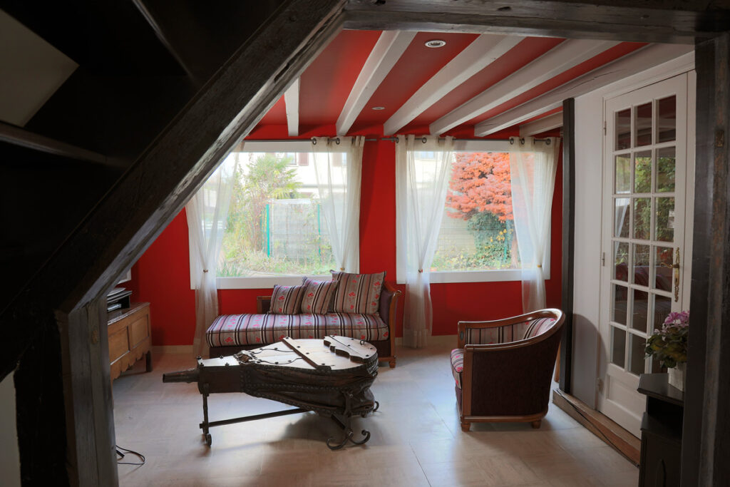 Volume unique pour cet intérieur, où les murs et le plafond rouge se confondent. L’espace est redéfini par l’effet soie sur la hotte et la couleur grise des poutres. Le jardin s’invite comme un tableau vivant dans ce salon